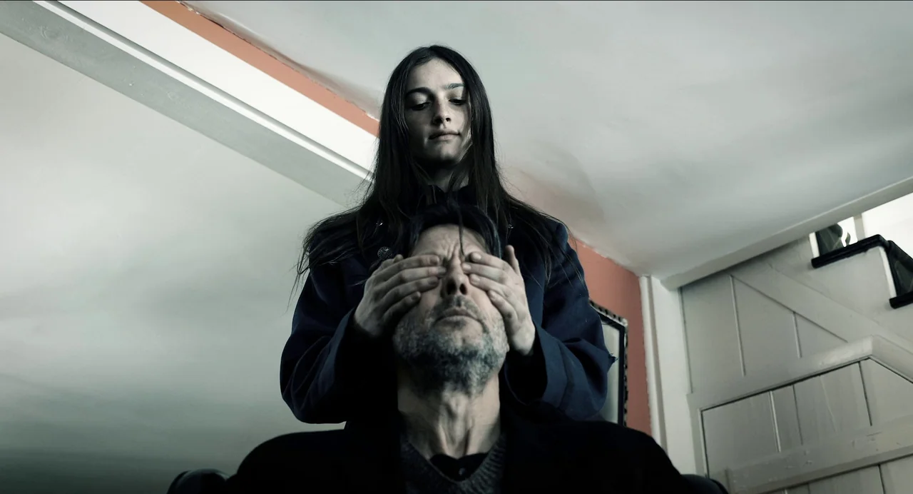Where The Devil Roams (2023) movie still: medium — A stairwell with white-painted walls and a peach-colored accent strip near the ceiling. A woman…
