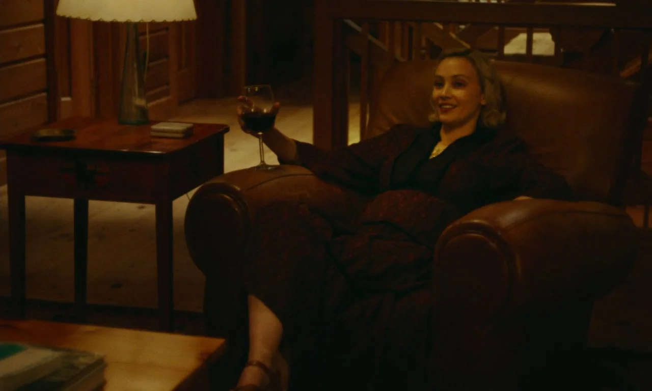Black Bear (2020) movie still: medium — A woman relaxes in a dark, warm-toned living room with wood paneling. She is sitting in a large leather…