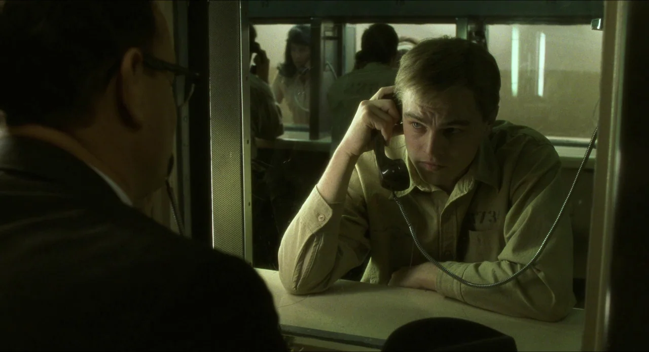 Catch Me If You Can (2002) movie still: medium — A sterile visiting area features a thick glass partition separating two people. A young man sits at…