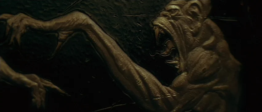 Planet of the Apes (2001) movie still: closeup — A dark stone carving mounted on a wall shows an aggressive mythical creature. The creature has…