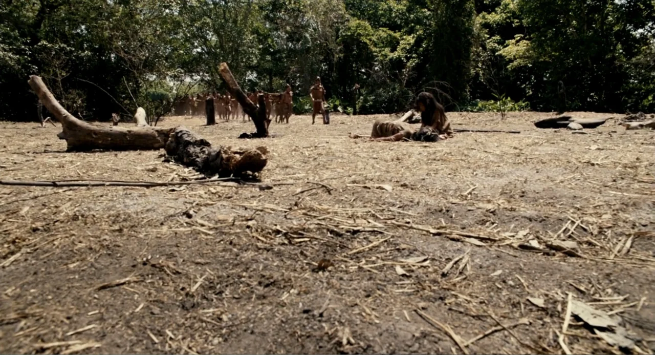 Apocalypto (2006) movie still: wide — A clearing in a dense tropical forest with dry soil and scattered dead wood. A person kneels beside another…