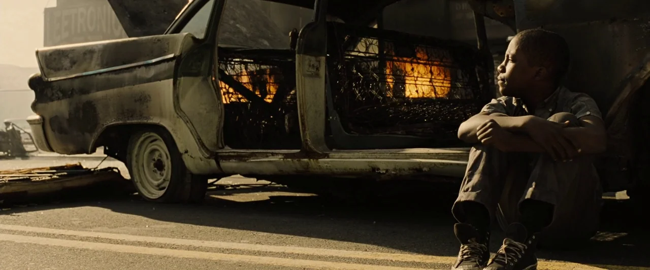 Across the Universe (2007) movie still: medium — A charred car frame burns intensely on a street littered with debris. A young boy sits on the…