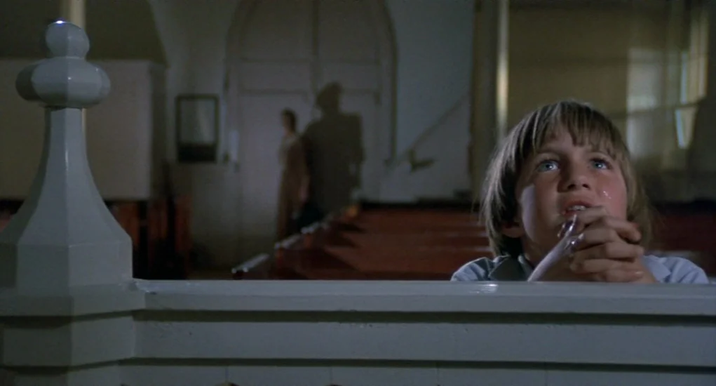 The Other (1972) movie still: medium — A dimly lit church interior with rows of wooden benches in the background. A young boy rests his arms on the…