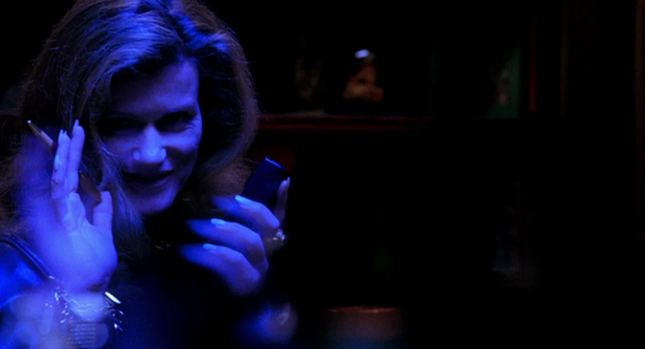 Office Killer (1997) movie still: closeup — A dimly lit space is bathed in intense blue light from a nearby source. A woman with light hair gestures…