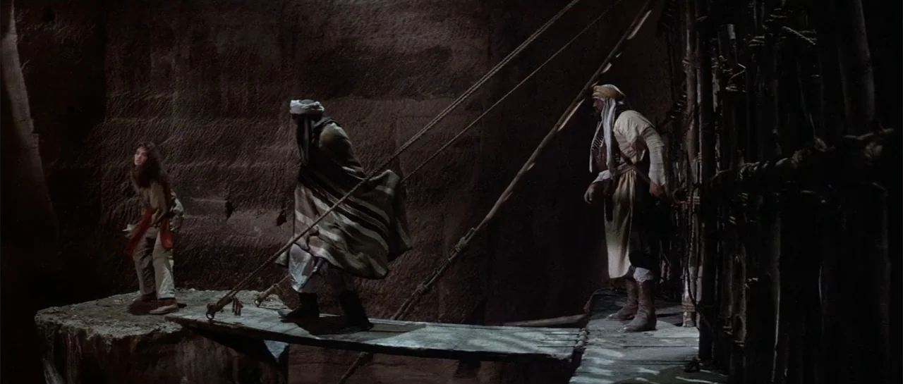 Sahara (1995) movie still: wide — A dark subterranean stone cavern features a narrow wooden bridge crossing a deep void. Three people in 1930s-style…