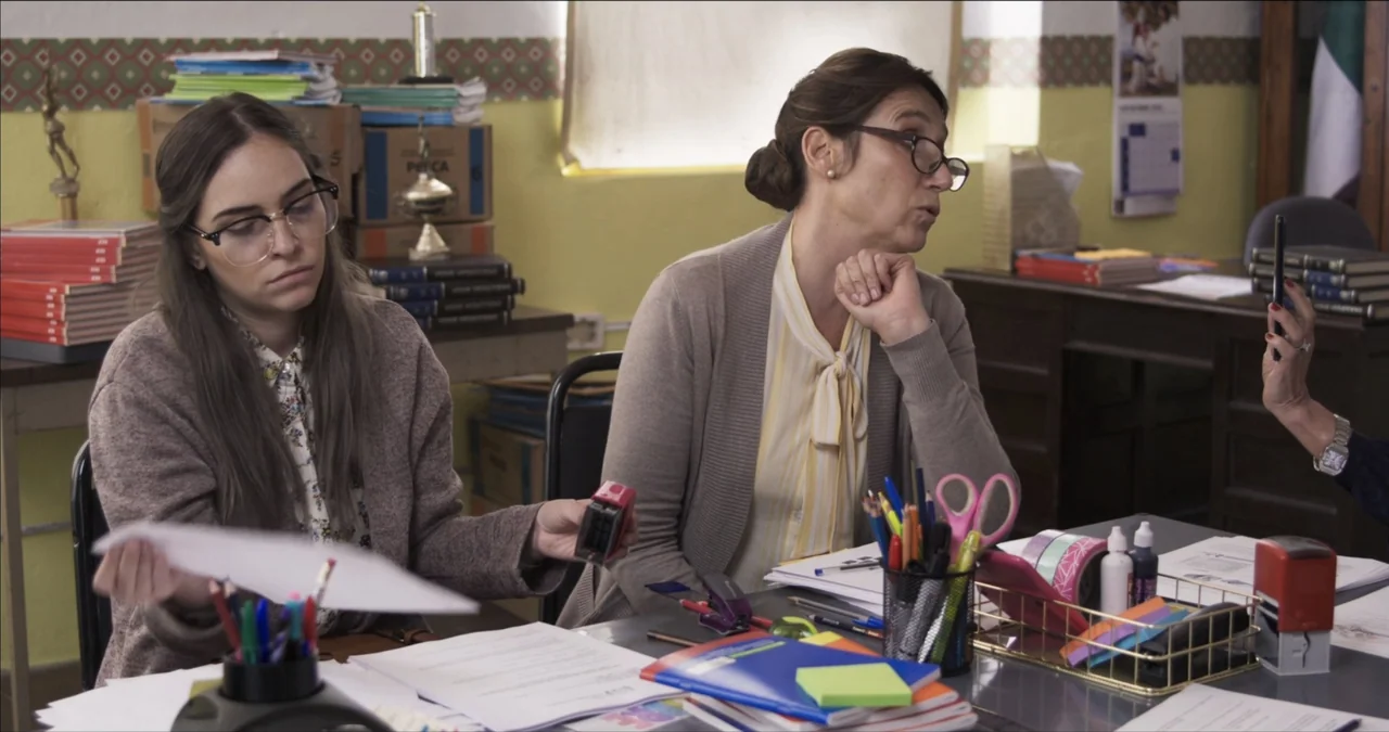 Bendita Suegra (2023) movie still: medium — An office filled with paperwork, cardboard boxes, and various desk supplies on flat surfaces. Two women…