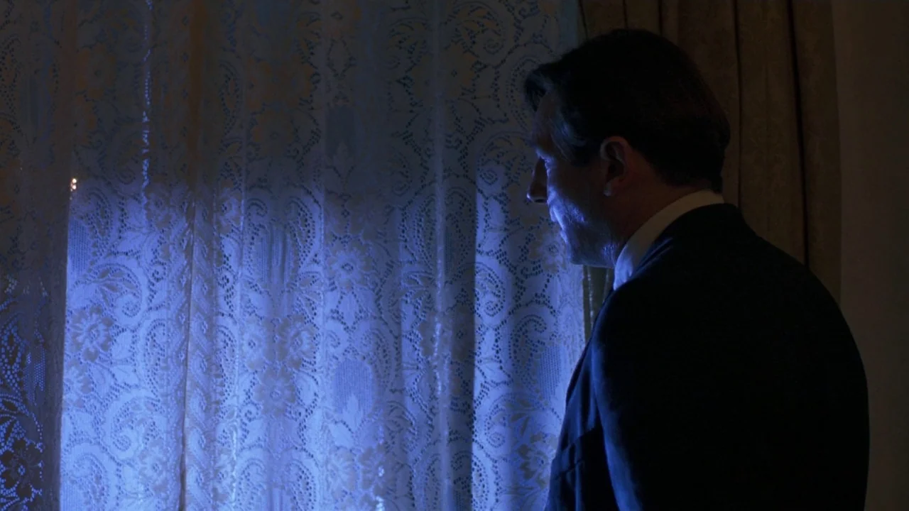 Michael Collins (1996) movie still: medium — A dark indoor space with white lace curtains covering a window. A man in a dark suit stands looking out…