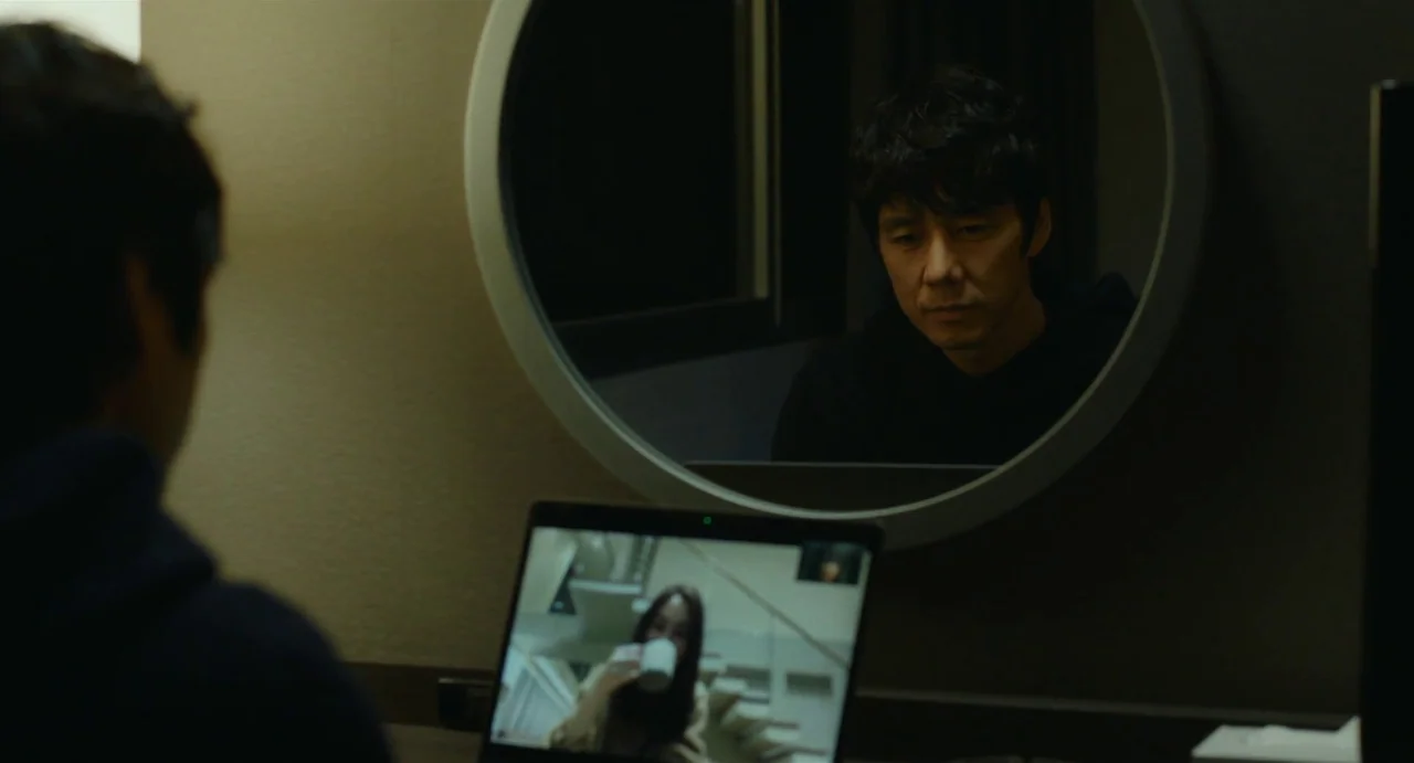 Drive My Car (2021) movie still: medium — A dimly lit room features a man reflected in a large circular wall mirror. He sits before a laptop…