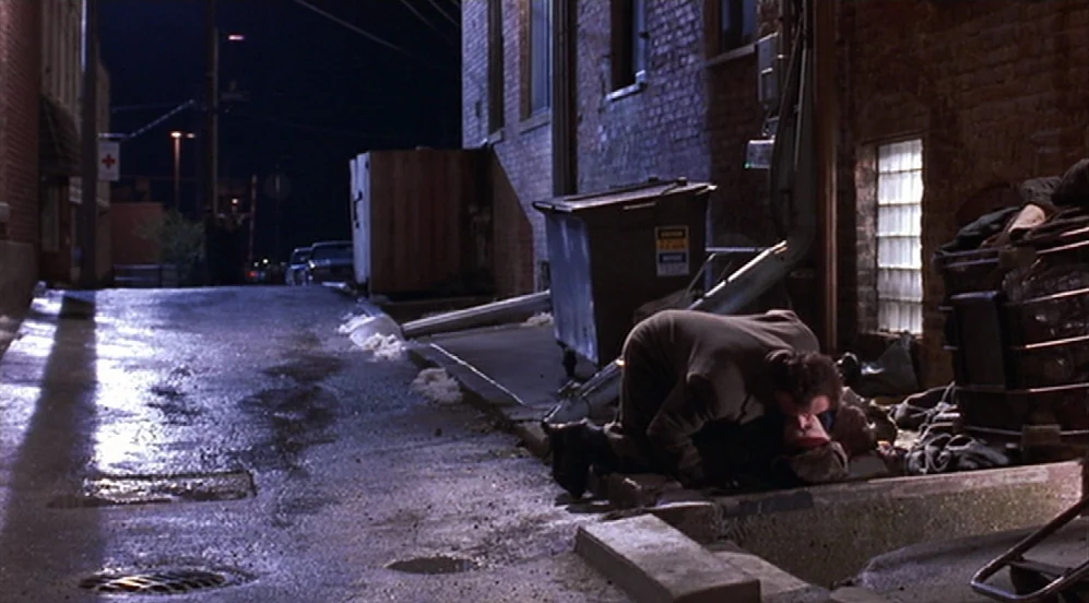 Groundhog Day (1993) movie still: wide — A narrow brick urban alleyway features a metal dumpster and wet pavement at night. One man in a trench coat…