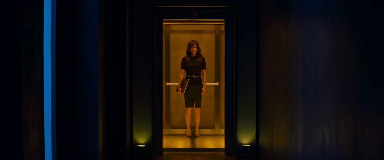 Elizabeth Harvest movie still: fullbody — A woman stands centered inside a metallic elevator framed by a dark, dim corridor. The elevator interior…