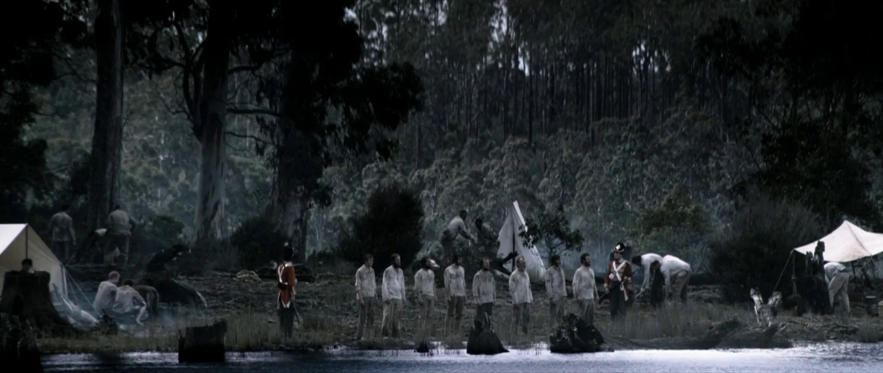 Van Diemens Land (2009) movie still: wide — A group of men in white shirts stand at the edge of a lake. Several canvas tents are pitched near large…