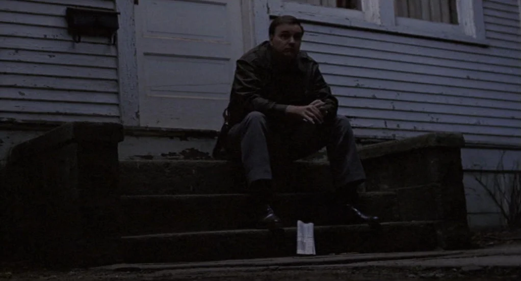 The Indian Runner (1991) movie still: fullbody — A man sits on the dark, concrete steps of a pale-painted suburban house. He rests with his hands…
