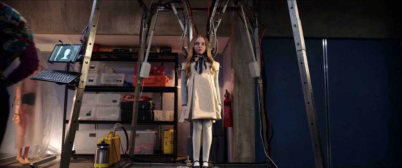 M3gan (2022) movie still: medium — A sterile laboratory setting contains metal structures, storage shelving with equipment, and cabling. A…