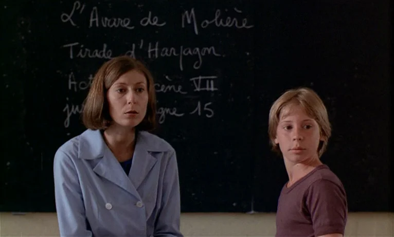 Small Change (1976) movie still: medium — A dark chalkboard with handwritten French script serves as the background for two people. A woman and a…
