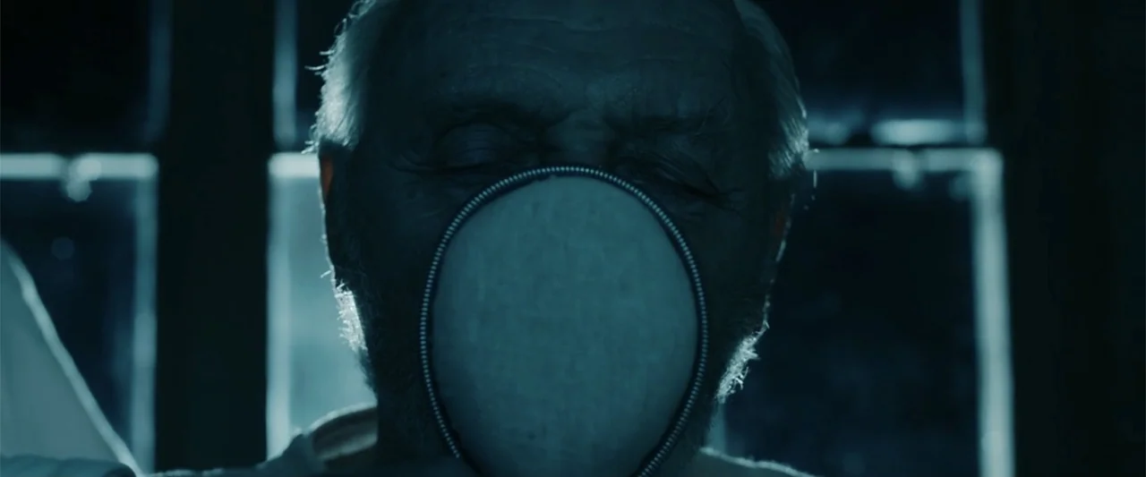 Freud's Last Session (2023) movie still: closeup — An interior space is dimly lit by a cold light source from behind. An elderly man wears a zip-up…