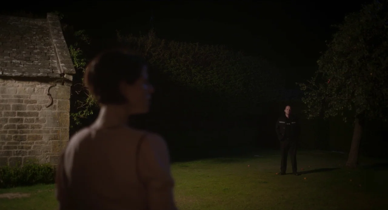 Men (2022) movie still: medium — A dark outdoor space features a stone wall structure on the left side. A man stands at a distance in the shadows…