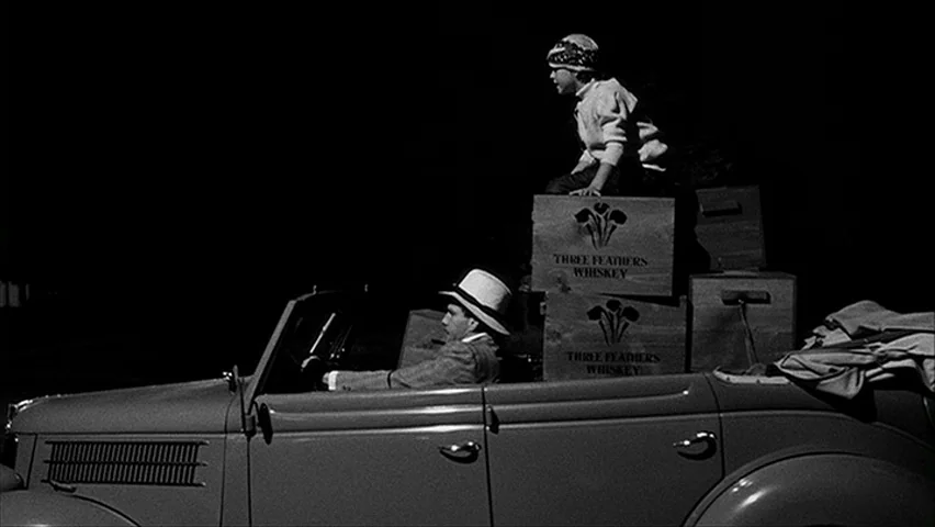Paper Moon (1971) movie still: medium — A man drives a 1930s convertible at night with a child perched on cargo. Wooden whiskey crates are stacked…
