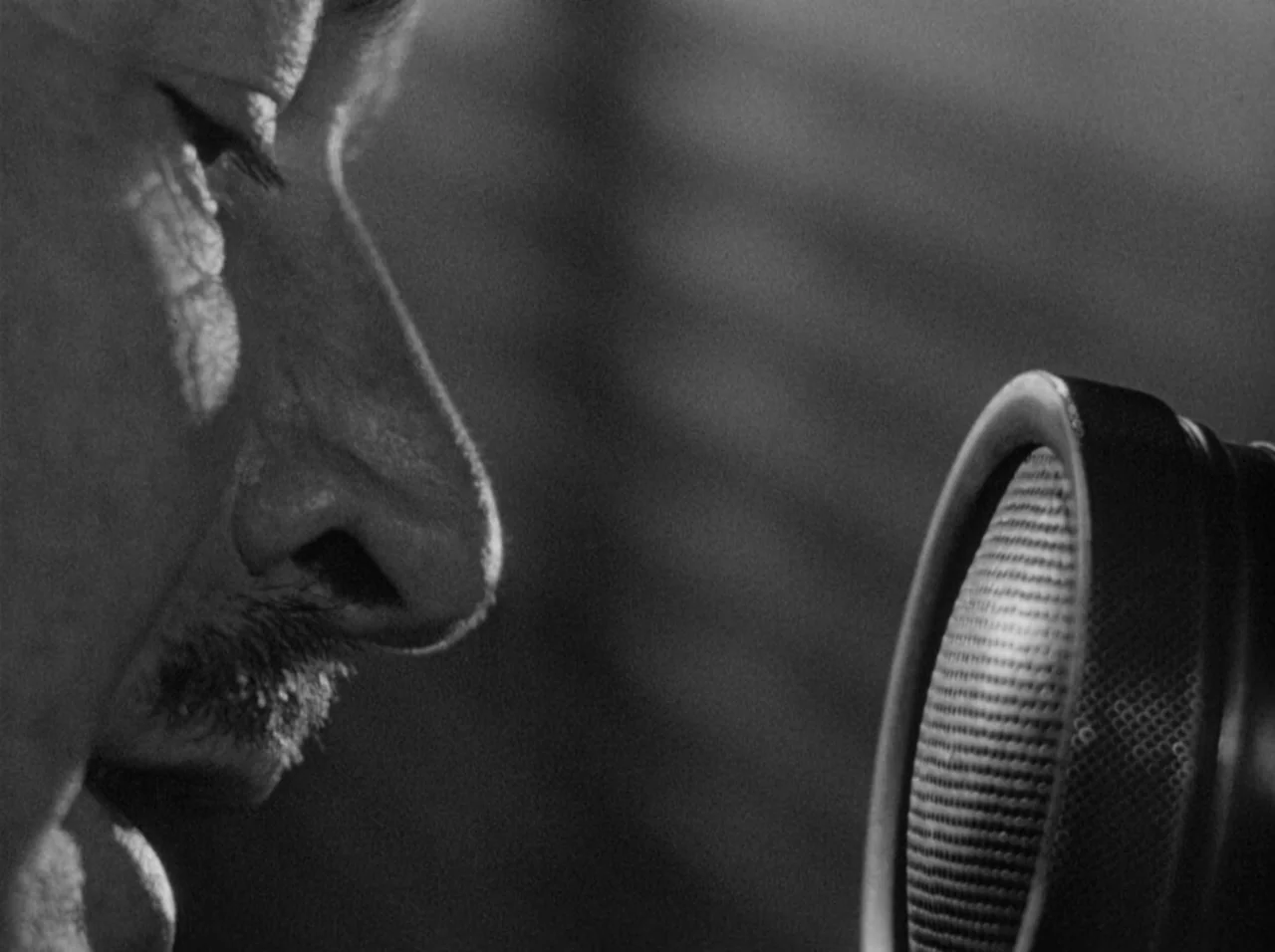 Gun Crazy (1950) movie still: closeup — A man speaks into a vintage microphone in a darkened recording space. Sharp shadows highlight the contours…