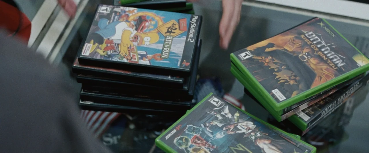 Roofman (2025) movie still: medium — A pile of video game cases rests on a glass surface. A person reaches toward the gaming titles from the left…