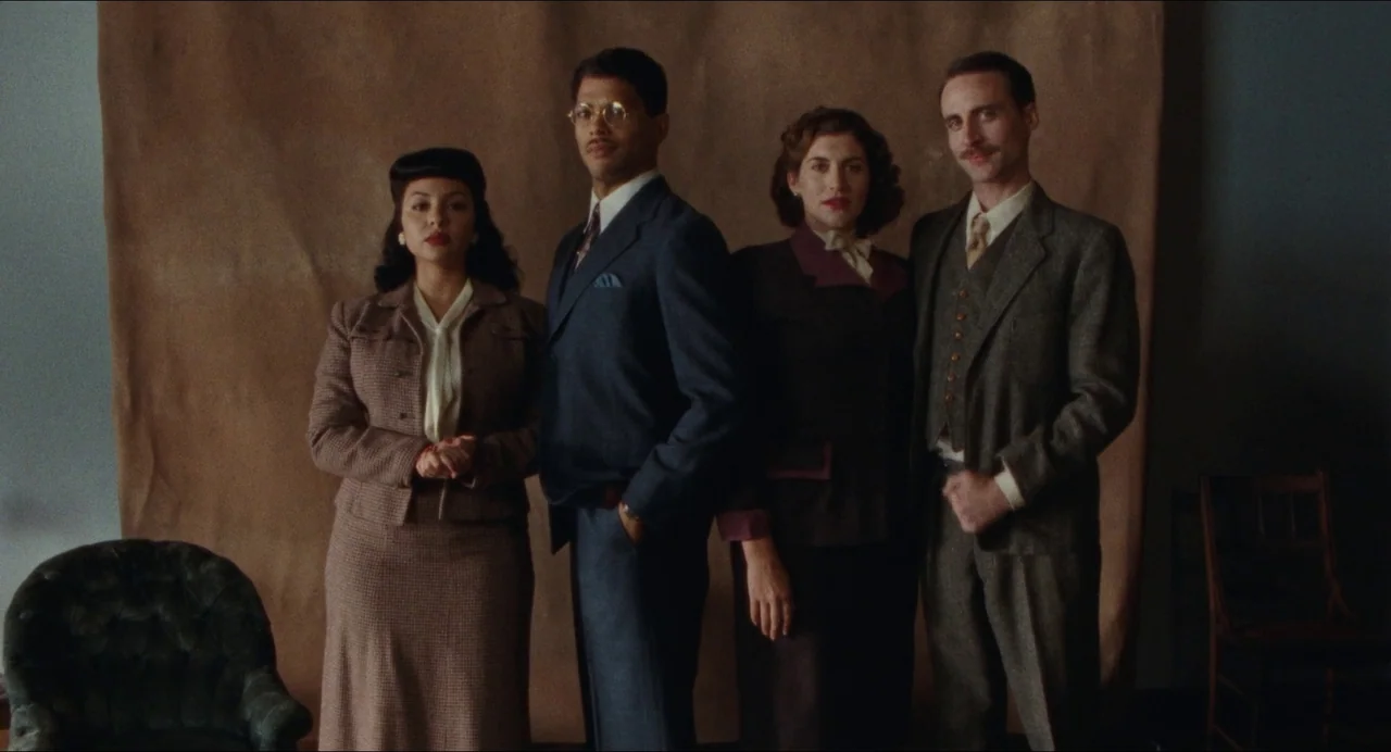 Origin (2023) movie still: fullbody — Four people stand side by side before a large, plain tan backdrop. They pose formally for a group photograph…