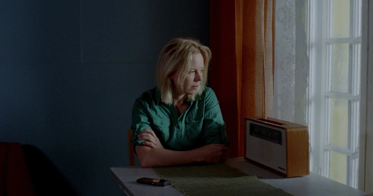 Fallen Leaves (2023) movie still: medium — A small dining area features a table beside a window with orange curtains. A blonde woman sits at the…
