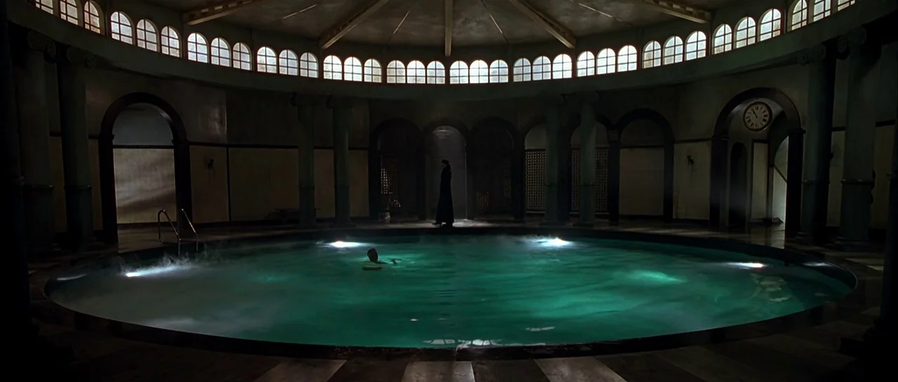 Dark City (1998) movie still: wide — A circular indoor pool hall features arched walls and high windows beneath a domed roof. One man floats in the…