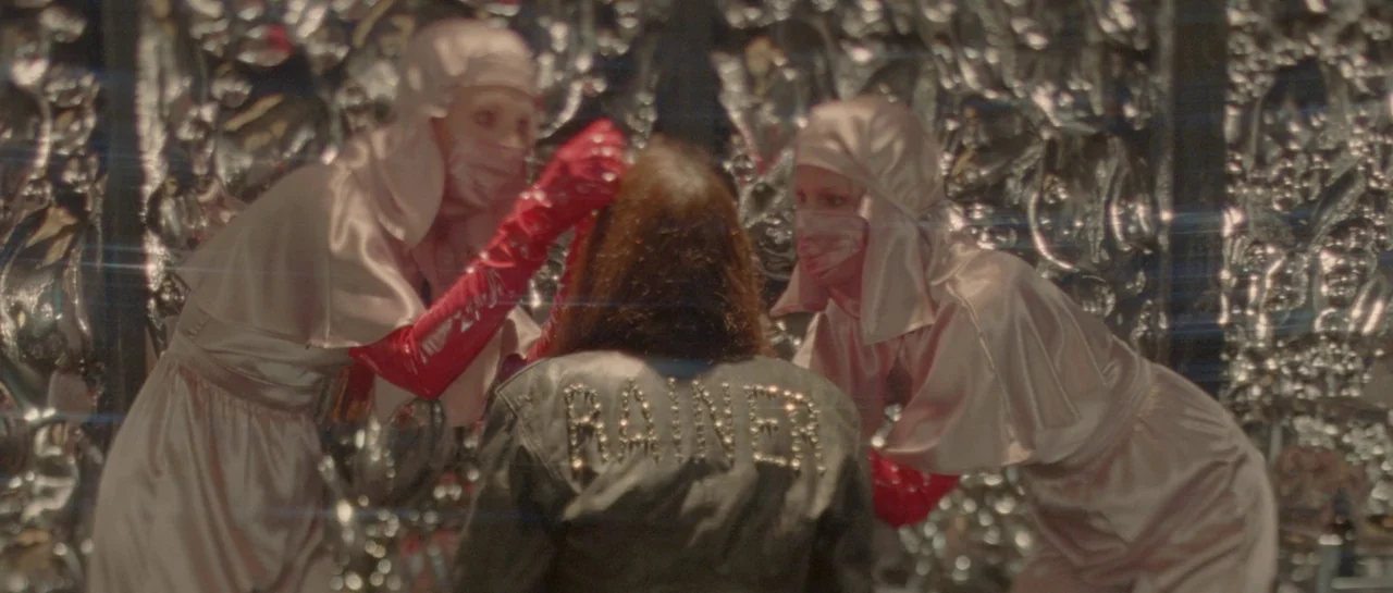 She Is Conann (2023) movie still: medium — An interior space features a backdrop of reflective, textured silver metallic surfaces. Two masked…