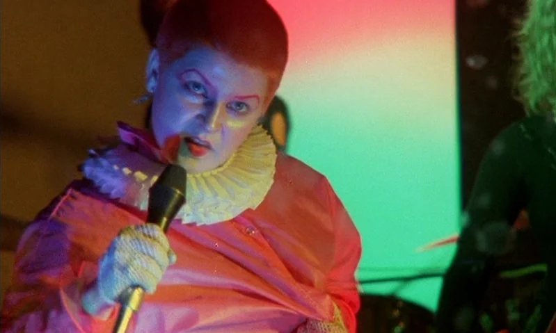Jubilee (1978) movie still: medium — A performer stands on a stage with neon red and green lights behind them. The individual sings into a black…