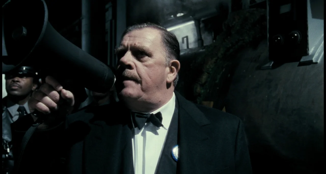 Batman (1989) movie still: medium — A dark night street setting features a man in formal wear. The man holds a black megaphone to his mouth to…