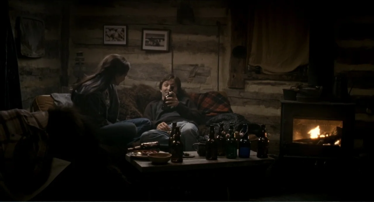 Colin Quinn: Red State, Blue State (2019) movie still: medium — A rustic interior of a log cabin with a wood stove and log walls. A man and a woman…