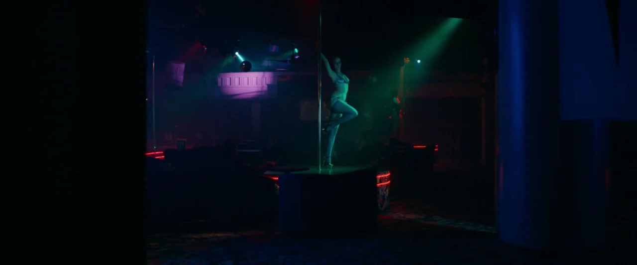 Little Woods (2018) movie still: fullbody — A dark night club features a woman dancing on a center stage pole. Green and red neon spotlights…