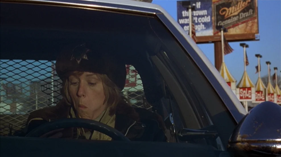 Fargo (1996) movie still: closeup — A woman in a police uniform sits in the driver seat of a patrol car. She is chewing while looking ahead through…