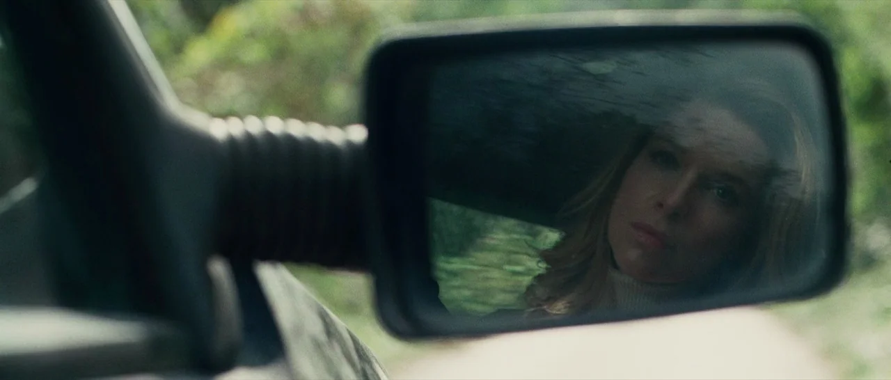 The American (2010) movie still: closeup — A vehicle side mirror reflects a woman looking ahead against a blurred green background. The mirror…