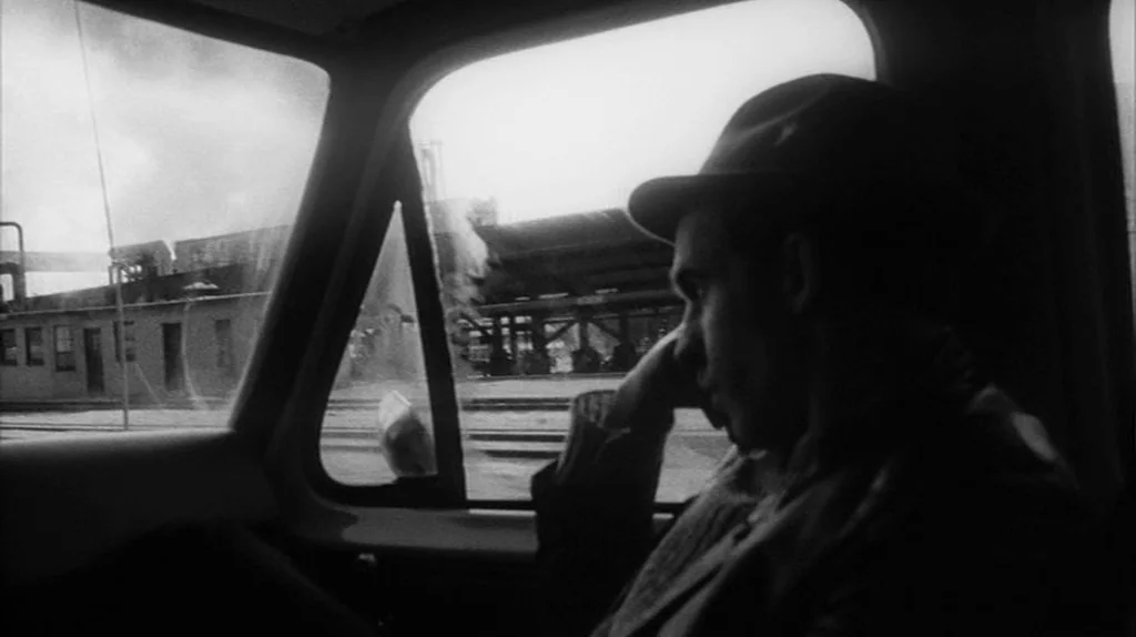 Stranger Than Paradise (1984) movie still: medium — The interior of a moving car is captured with view of industrial buildings outside. A man in a…