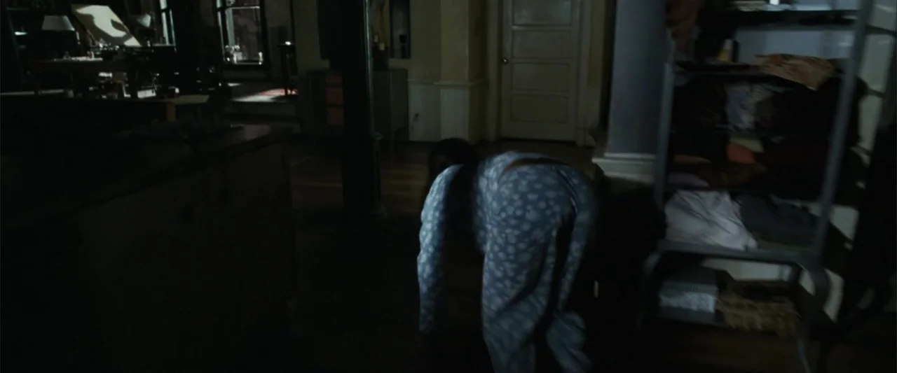 Catwoman (2004) movie still: medium — An dimly lit apartment interior with wooden flooring and a metal storage shelf. A woman in floral pajamas…