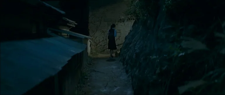 Mother (2009) movie still: wide — A narrow dark alleyway is flanked by a stone retaining wall and building roof. A lone schoolgirl walks away from…