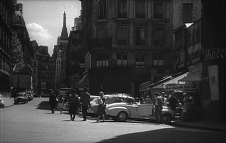 Le Signe du Lion (1962) movie still: wide — A Paris street intersection features multi-story stone buildings with tall rectangular windows and…