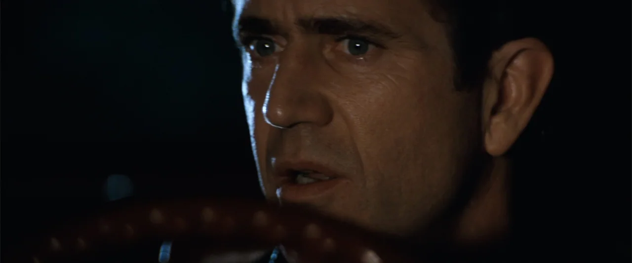 Conspiracy Theory (1997) movie still: closeup — The interior of a vehicle is dark with limited visibility. A man in his forties holds the steering…