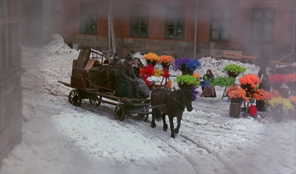 Fanny and Alexander (1982) movie still: wide — A snow-covered public square features a brick building facade and rows of colorful flower displays. A…