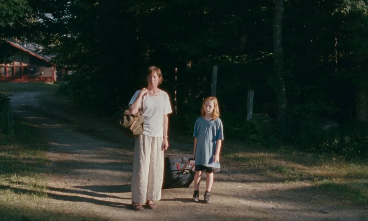 Janet Planet (2023) movie still: fullbody — An adult woman and a young girl stand together on a dirt forest road. They carry bags while positioned…
