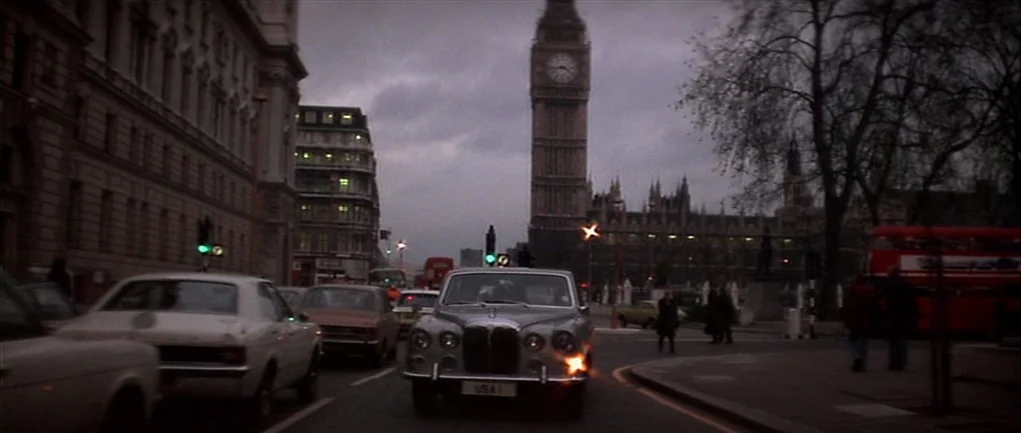 The Omen (1976) movie still: wide — An urban London street intersection features Big Ben standing tall against a grey sky. Traffic flows steadily…