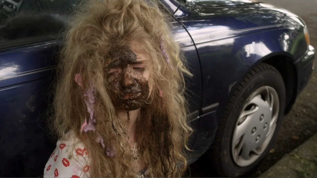 Jinxed (2013) movie still: medium — A person sits on the ground next to a dark blue sedan on a paved street. The subject has long blonde hair and is…