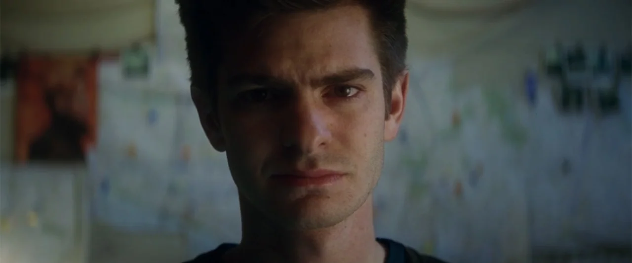 The Amazing Spider-Man 2 (2014) movie still: closeup — An interior space with a blurred wall featuring pinned notes or maps behind him. A young man…