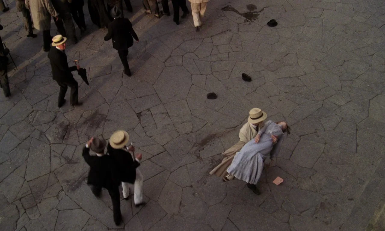 A Room With A View (1985) movie still: wide — A wide stone-paved plaza populated by men in early twentieth-century formal attire. A man lifts a…