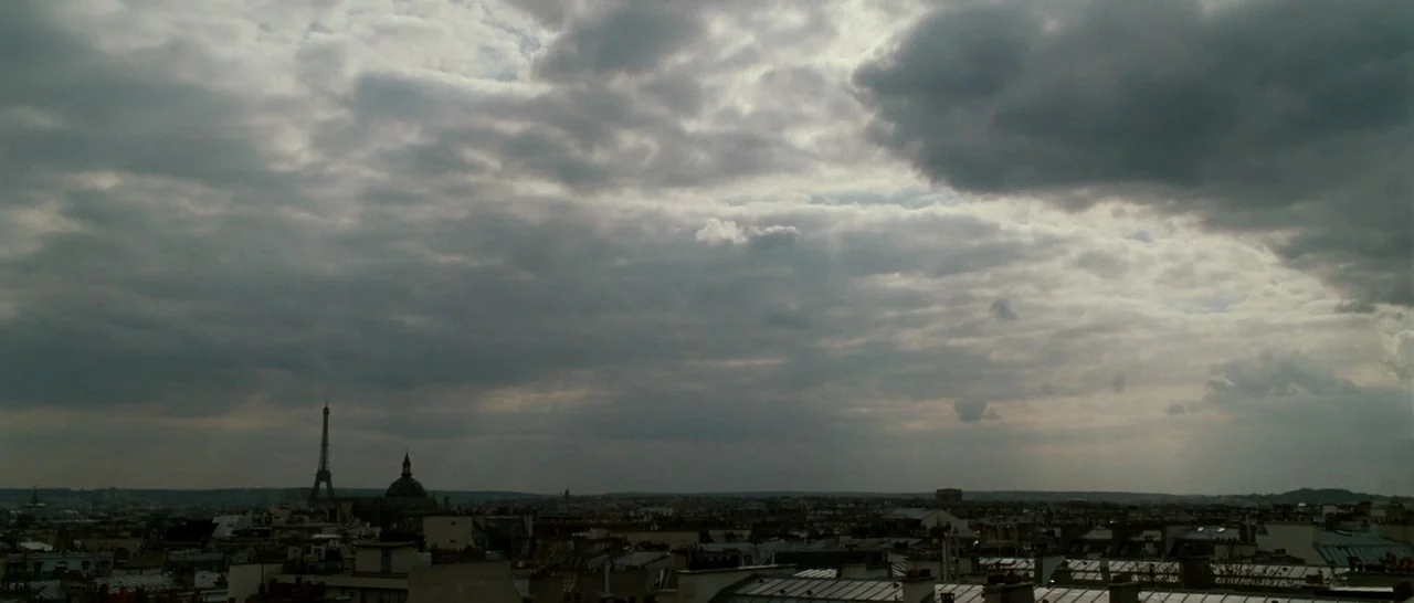 La Vie En Rose (2007) movie still: establishing — A wide view of the Paris skyline shows rooftops under a heavy overcast sky. The Eiffel Tower is…
