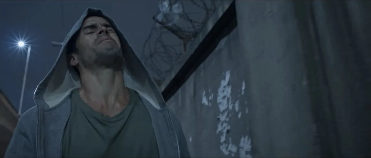 Gunman (2025) movie still: medium — A narrow urban alleyway at night features a concrete wall topped with barbed wire. A man in a hoodie stands…
