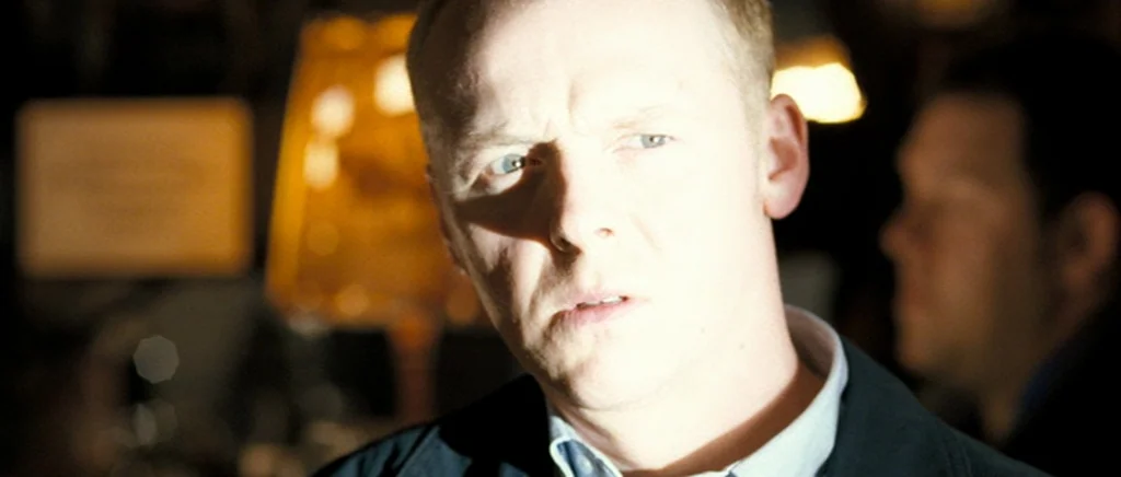 Hot Fuzz (2007) movie still: closeup — A dimly lit indoor space with blurred background elements suggesting a social gathering area. A man stands in…