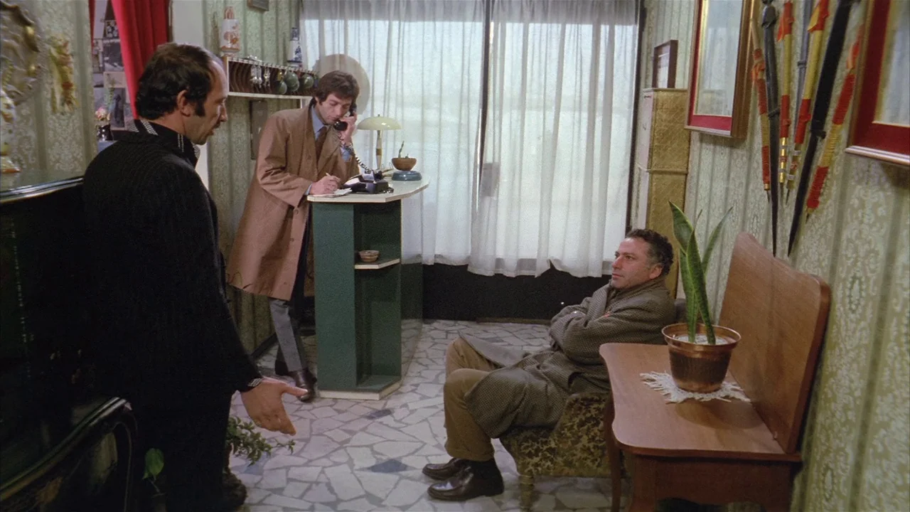 Death Occurred Last Night (1970) movie still: wide — A small reception area features wallpaper with a vintage pattern and a stone floor. Three men…