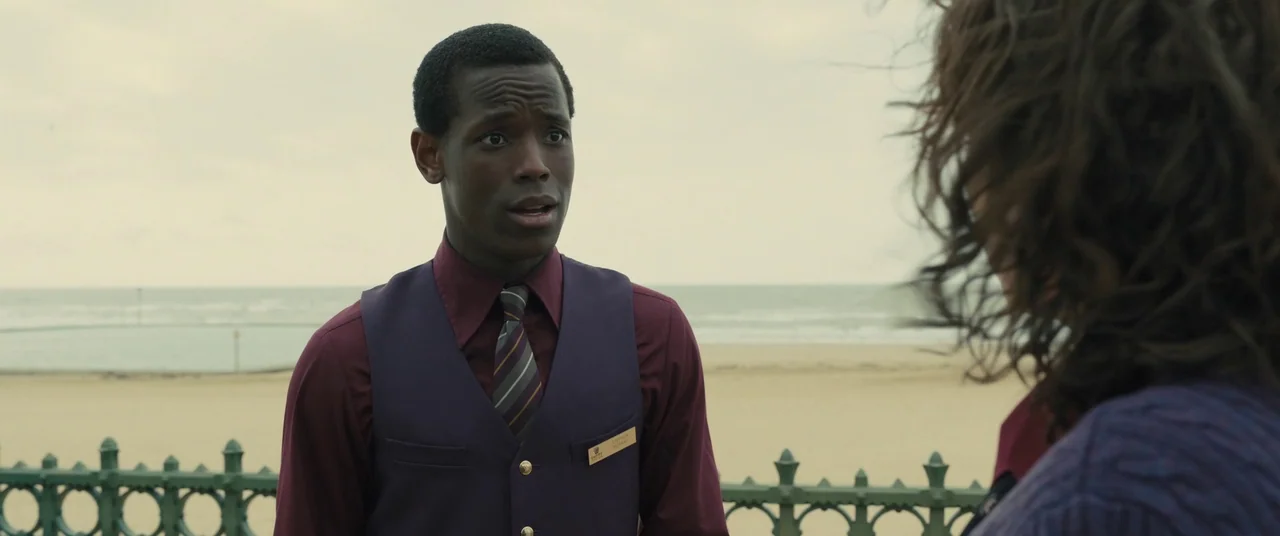 Empire of Light (2022) movie still: medium — An overcast promenade overlooking a sandy beach with a cast-iron fence. A young man in a uniform stands…