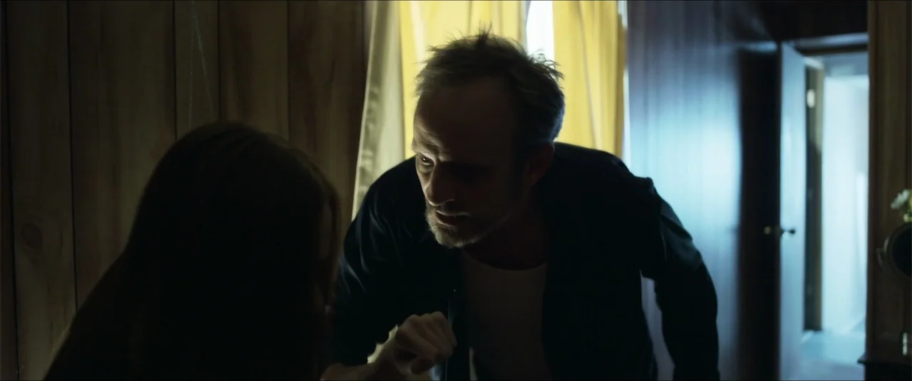 Captive (2020) movie still: medium — A dark bedroom featuring wood-paneled walls and a window with yellow curtains. A man leans forward toward…